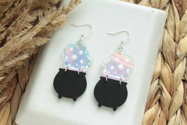 May include: A pair of iridescent earrings featuring a black cauldron with a swirling cloud of stars above it.