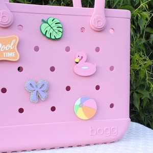 May include: A light pink rubber tote bag with a perforated design and two handles. The bag is decorated with five colorful charms: a green monstera leaf, a pink flamingo, a glittery purple bow, a beach ball, and a sign reading IT'S POOL TIME.