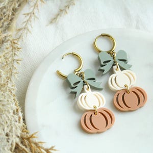 May include: A pair of fall-themed earrings featuring gold-colored hoops, sage green bows, and three stacked pumpkin shapes in cream and terracotta colors. The earrings are displayed on a white marble plate.