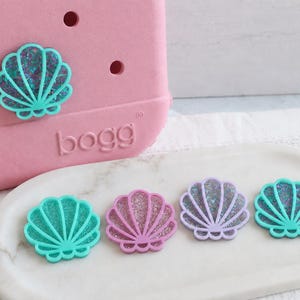 May include: A pink Bogg bag with a teal seashell charm attached, alongside four additional seashell charms in teal, pink, lavender, and teal, each featuring a glittery interior, arranged on a white marble-patterned oval tray.