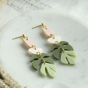 May include: A pair of dangle earrings with a pink, white, and green color scheme. The earrings feature three geometric shapes and two leaf-shaped charms.