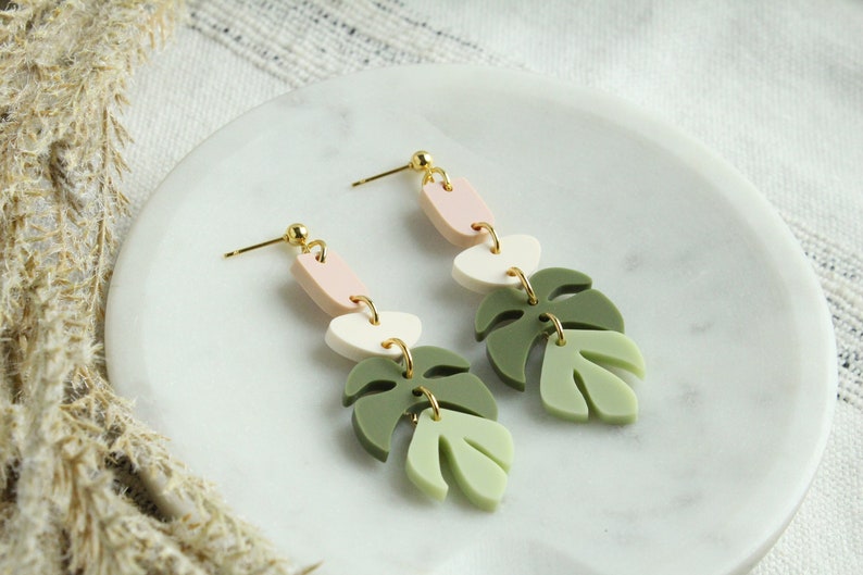 May include: A pair of dangle earrings with a pink, white, and green color scheme. The earrings feature three geometric shapes and two leaf-shaped charms.