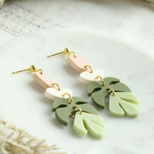 May include: A pair of dangle earrings with three layered polymer clay shapes. The top shape is a light pink oval, the middle shape is a white oval, and the bottom shape is a green leaf. The earrings have gold hooks.