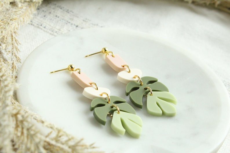 May include: A pair of dangle earrings with three layered polymer clay shapes. The top shape is a light pink oval, the middle shape is a white oval, and the bottom shape is a green leaf. The earrings have gold hooks.