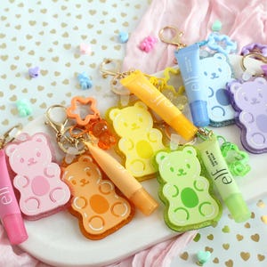 May include: A collection of colorful gummy bear-shaped keychains, each paired with a matching e.l.f. Squeeze Me lip balm tube. The keychains feature translucent bear charms in pink, orange, yellow, green, blue, and purple, attached to gold-tone metal rings with small star or bear-shaped accents.
