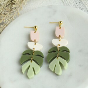 May include: A pair of dangle earrings with a layered design. The earrings feature three geometric shapes in pink, white, and green, followed by a green leaf shape. The earrings are made of polymer clay and have gold-toned hardware.