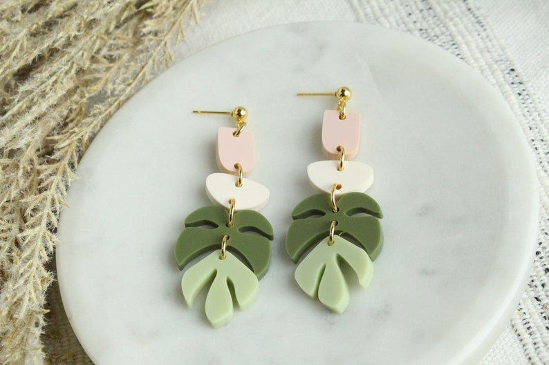 May include: A pair of dangle earrings with a layered design. The earrings feature three geometric shapes in pink, white, and green, followed by a green leaf shape. The earrings are made of polymer clay and have gold-toned hardware.