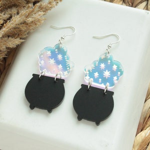 May include: A pair of black cauldron earrings with iridescent blue and pink clouds of smoke with white stars. The earrings are made of acrylic and have silver hooks.