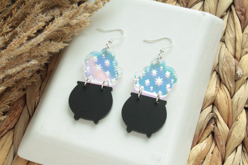 May include: A pair of black cauldron earrings with iridescent blue and pink clouds of smoke with white stars. The earrings are made of acrylic and have silver hooks.