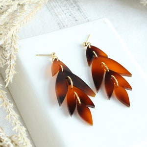 May include: A pair of long, brown and gold earrings with a leaf design. The earrings are made of multiple layers of brown leaves with gold accents.