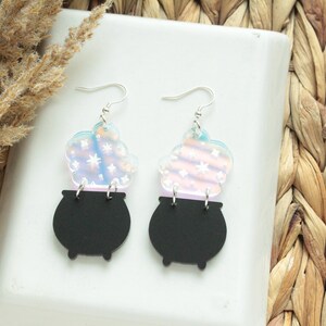 May include: A pair of iridescent earrings featuring a black cauldron with a swirling, iridescent cloud of smoke above it. The earrings are made of acrylic and have silver hooks.