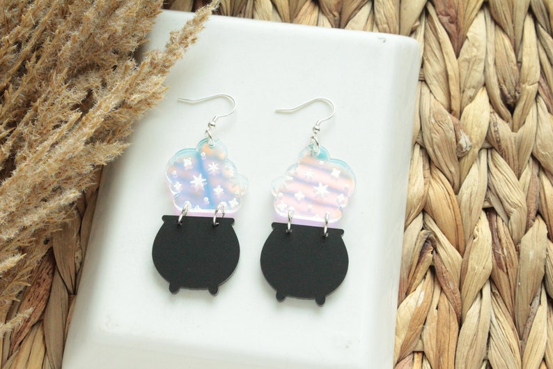 May include: A pair of iridescent earrings featuring a black cauldron with a swirling, iridescent cloud of smoke above it. The earrings are made of acrylic and have silver hooks.