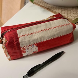 Könnte beinhalten: Eine rote gesteppte Stiftebox mit Reißverschluss und einem beigefarbenen Stoffpanel mit roten Nähten. Die Box ist mit einem Blumenmuster verziert und hat einen Stift daneben liegen.