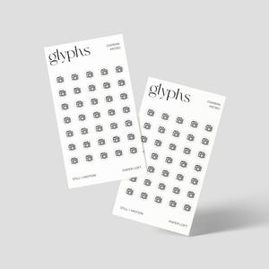 May include: Two white sticker sheets with a minimalist design. Each sheet features a grid of small, black camera icons. The word "glyphs" is at the top, with "camera micro" and "still + motion" text. The stickers are for Paper Loft.