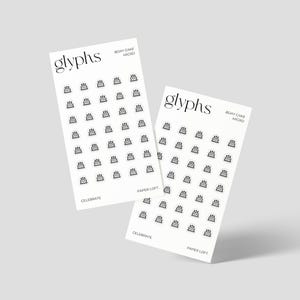 May include: Two white cards with the word "glyphs" in a stylish font. Each card has a grid of small black cake illustrations. The text "BIRTHDAY CAKE MICRO" and "CELEBRATE" are also printed on the cards. The cards are from "PAPER LOFT".