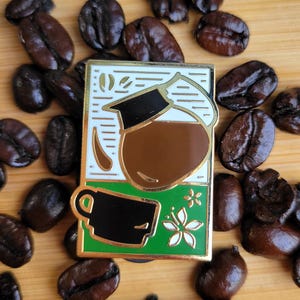 May include: A rectangular enamel pin featuring a graphic illustration of a glass coffee carafe filled with brown liquid above a black coffee cup. The design includes a white background with thin horizontal lines and a green base with a white flower.