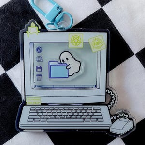 Peut inclure: Un porte-clés bleu clair avec un design d'ordinateur et un motif de fantôme sur l'écran. Le porte-clés a un fermoir bleu clair et comprend le texte "TECH SUPPORT 666-HELP". Le design de l'ordinateur comprend un clavier et une souris.