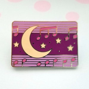 May include: A purple and pink enamel pin with a crescent moon, stars, and musical notes. The pin has a gold outline.