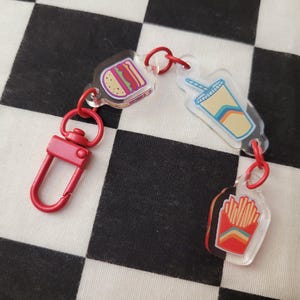 May include: A red keychain with a lobster clasp and three acrylic charms. The charms depict a hamburger, a soda cup, and a container of fries. The food-themed charms have colorful illustrations and are connected by red jump rings.