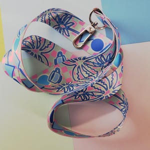 Op de afbeelding: Een roze, blauwe en groene lanyard met een palmboom- en bekerontwerp. De lanyard heeft een gouden metalen sluiting.