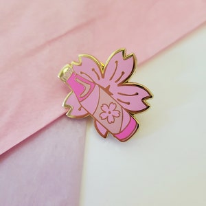 May include: A gold-tone enamel pin featuring a pink bottle with a cherry blossom design. The bottle has a pink and white striped pattern and a small cherry blossom on the side. The bottle is surrounded by a pink cherry blossom.