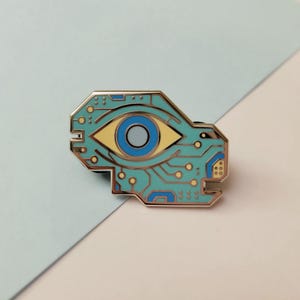 May include: A teal and gold enamel pin featuring an eye design within a circuit board outline. The eye has a yellow iris and a blue pupil. The pin is a unique accessory.