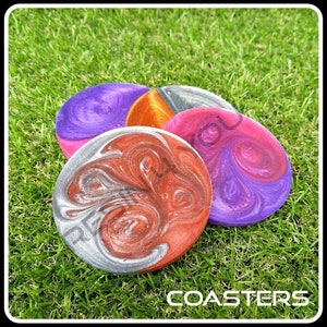 K&ouml;nnte beinhalten: Vier runde Untersetzer mit einem wirbelnden Muster aus silberner, roter, violetter und orangefarbener Farbe. Die Untersetzer befinden sich auf einer gr&uuml;nen Grasfl&auml;che. Das Wort "COASTERS" ist in wei&szlig;en Buchstaben auf dem Gras gedruckt.