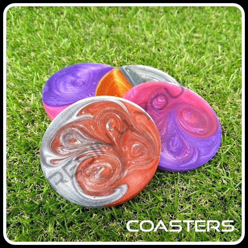 K&ouml;nnte beinhalten: Vier runde Untersetzer mit einem wirbelnden Muster aus silberner, roter, violetter und orangefarbener Farbe. Die Untersetzer befinden sich auf einer gr&uuml;nen Grasfl&auml;che. Das Wort "COASTERS" ist in wei&szlig;en Buchstaben auf dem Gras gedruckt.