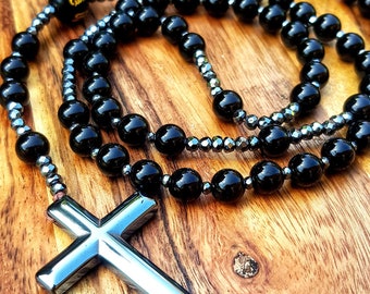 May include: A black onyx and hematite beaded necklace with a silver cross pendant. The necklace is lying on a wooden surface.