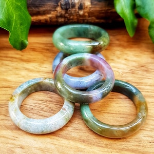 Puede incluir: Cuatro anillos de ágata verde, marrón y morado apilados sobre una superficie de madera. Los anillos están hechos de piedra pulida y tienen una forma redonda y lisa.