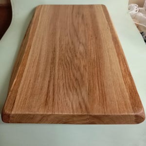 May include: A large, rectangular wooden cutting board with a natural wood finish. The board has a slightly rounded edge and is made of a single piece of wood.