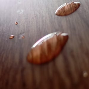 May include: Close-up of a wooden surface with two water droplets. The droplets are oval-shaped and reflect the wood grain.