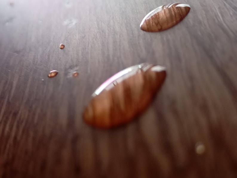 May include: Close-up of a wooden surface with two water droplets. The droplets are oval-shaped and reflect the wood grain.