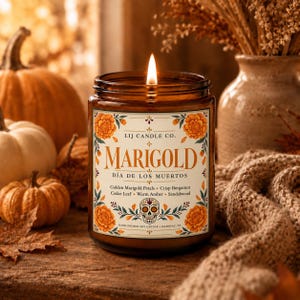 May include: A lit candle in an amber glass jar with a label reading LIJ CANDLE CO. MARIGOLD D&Iacute;A DE LOS MUERTOS. The label features orange marigold flowers, a skull illustration, and scent notes of Golden Marigold Petals, Crisp Bergamot, Cedar Leaf, Warm Amber, and Sandalwood.