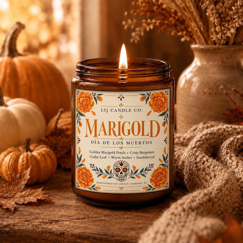 May include: A lit candle in an amber glass jar with a label reading LIJ CANDLE CO. MARIGOLD D&Iacute;A DE LOS MUERTOS. The label features orange marigold flowers, a skull illustration, and scent notes of Golden Marigold Petals, Crisp Bergamot, Cedar Leaf, Warm Amber, and Sandalwood.