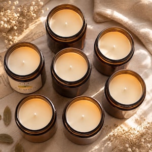 May include: Seven amber glass jars filled with cream-colored wax candles, each featuring a single centered wick. One jar displays a white label with the text "LEMON" visible. The candles are arranged in a cluster, showcasing the smooth wax surface.