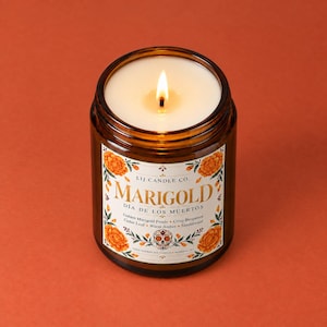 May include: A lit soy candle in an amber glass jar with a white label featuring orange marigold flowers, a decorative skull, and the text LIJ CANDLE CO. MARIGOLD D&Iacute;A DE LOS MUERTOS, listing scent notes of golden marigold petals and sandalwood.