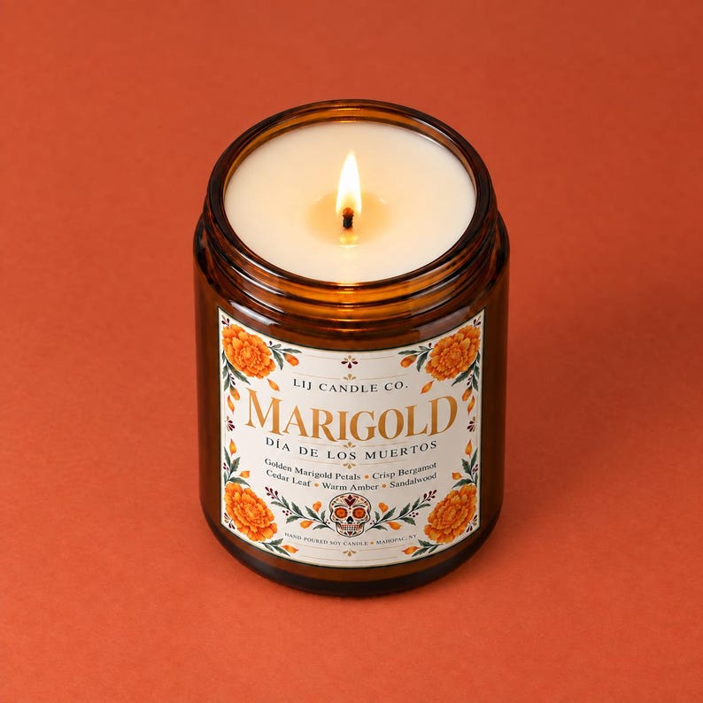 May include: A lit soy candle in an amber glass jar with a white label featuring orange marigold flowers, a decorative skull, and the text LIJ CANDLE CO. MARIGOLD D&Iacute;A DE LOS MUERTOS, listing scent notes of golden marigold petals and sandalwood.