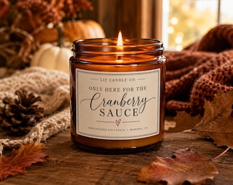 May include: A lit soy candle in an amber glass jar with a white label that reads LIJ CANDLE CO. ONLY HERE FOR THE Cranberry SAUCE with a small heart icon and the text HAND-POURED SOY CANDLE • MAHOPAC, NY, featuring a single burning wick.