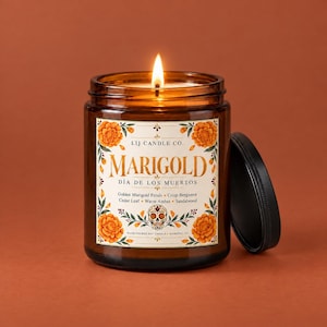 May include: A lit soy candle in an amber glass jar with a black lid. The label features orange marigold flowers, a skull illustration, and the text: LIJ CANDLE CO. MARIGOLD D&Iacute;A DE LOS MUERTOS Golden Marigold Petals, Crisp Bergamot, Cedar Leaf, Warm Amber, Sandalwood.