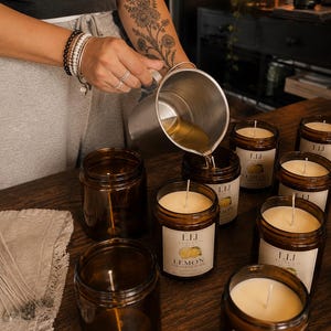 May include: A metal pouring pitcher filled with melted wax being poured into amber glass candle jars. Each jar features a white label with the text LIJ CANDLE CO and LEMON SUGARED COOKIE, containing a centered wick and off-white wax.