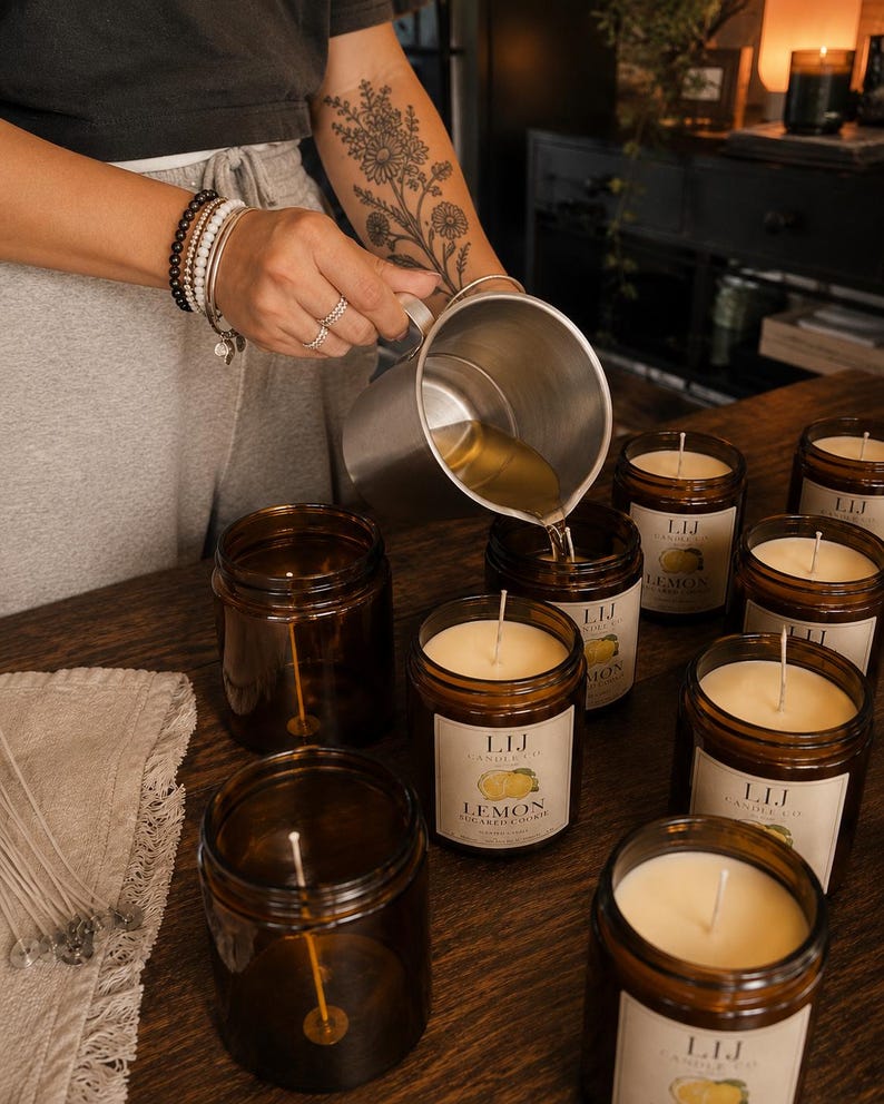 May include: A metal pouring pitcher filled with melted wax being poured into amber glass candle jars. Each jar features a white label with the text LIJ CANDLE CO and LEMON SUGARED COOKIE, containing a centered wick and off-white wax.