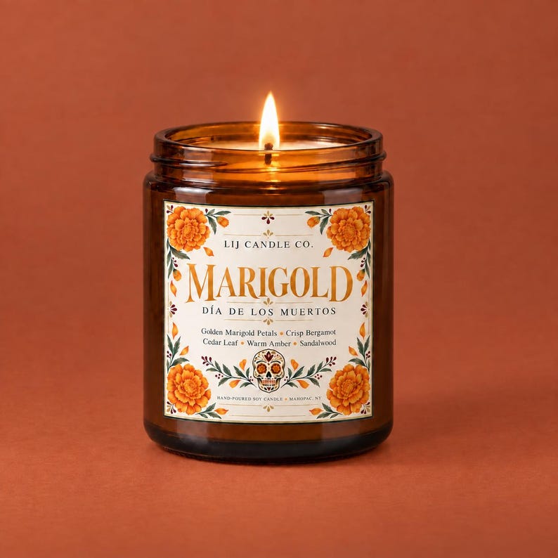 May include: A lit soy candle in a brown glass jar with a white label featuring orange marigold flowers and a central skull illustration. The text reads LIJ CANDLE CO. MARIGOLD D&Iacute;A DE LOS MUERTOS Golden Marigold Petals, Crisp Bergamot, Cedar Leaf, Warm Amber, Sandalwood.