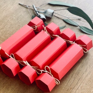 May include: A set of eight red paper Christmas crackers with white string ties. The crackers are arranged in a fan shape, with the ends of the crackers pointing towards the centre.