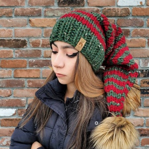 Könnte beinhalten: Eine grün-rot gestreifte Strickmütze mit einem langen, schlaffen Rand und einem Bommel aus Kunstfell. Die Mütze hat ein braunes Leder-Etikett mit dem Text "The Knit Wit" darauf.