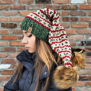 May include: A green, red and white striped knitted hat with a long, pointed top and a faux fur pom-pom. The hat is being worn by a person with long brown hair.