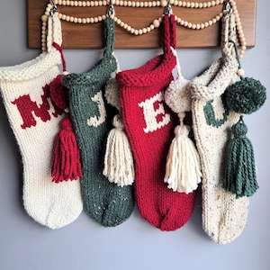 May include: Four knitted Christmas stockings with pom-poms and tassels hanging on a wooden rack. The stockings are white, green, red, and cream with the letters "MERRY" knitted on them.