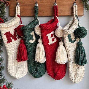 May include: Four hand-knit Christmas stockings hanging from a wooden rack. The stockings are cream, dark green, and red, featuring knit letters M, J, E, and U. Each stocking is decorated with yarn pom-poms and tassels in coordinating colors.