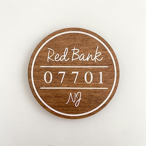 Op de afbeelding: Een rond houten bord met een witte rand en witte tekst. Het bord zegt "Red Bank 07701 NJ".