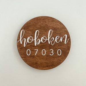 Op de afbeelding: Een rond houten bord met de tekst "hoboken 07030" in witte verf.
